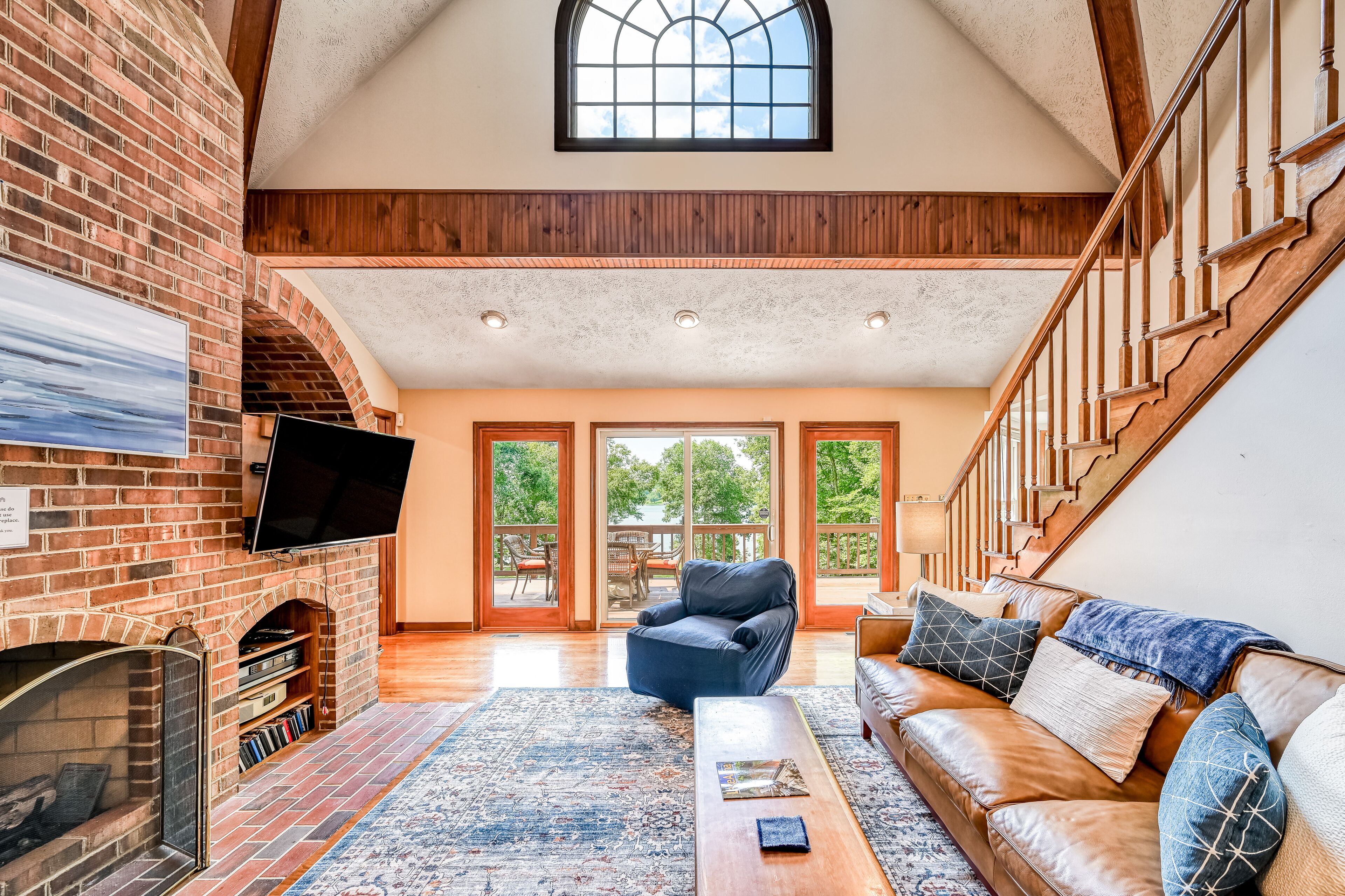 A spacious living room with a brick fireplace, a leather sofa, and a large window overlooking a deck.