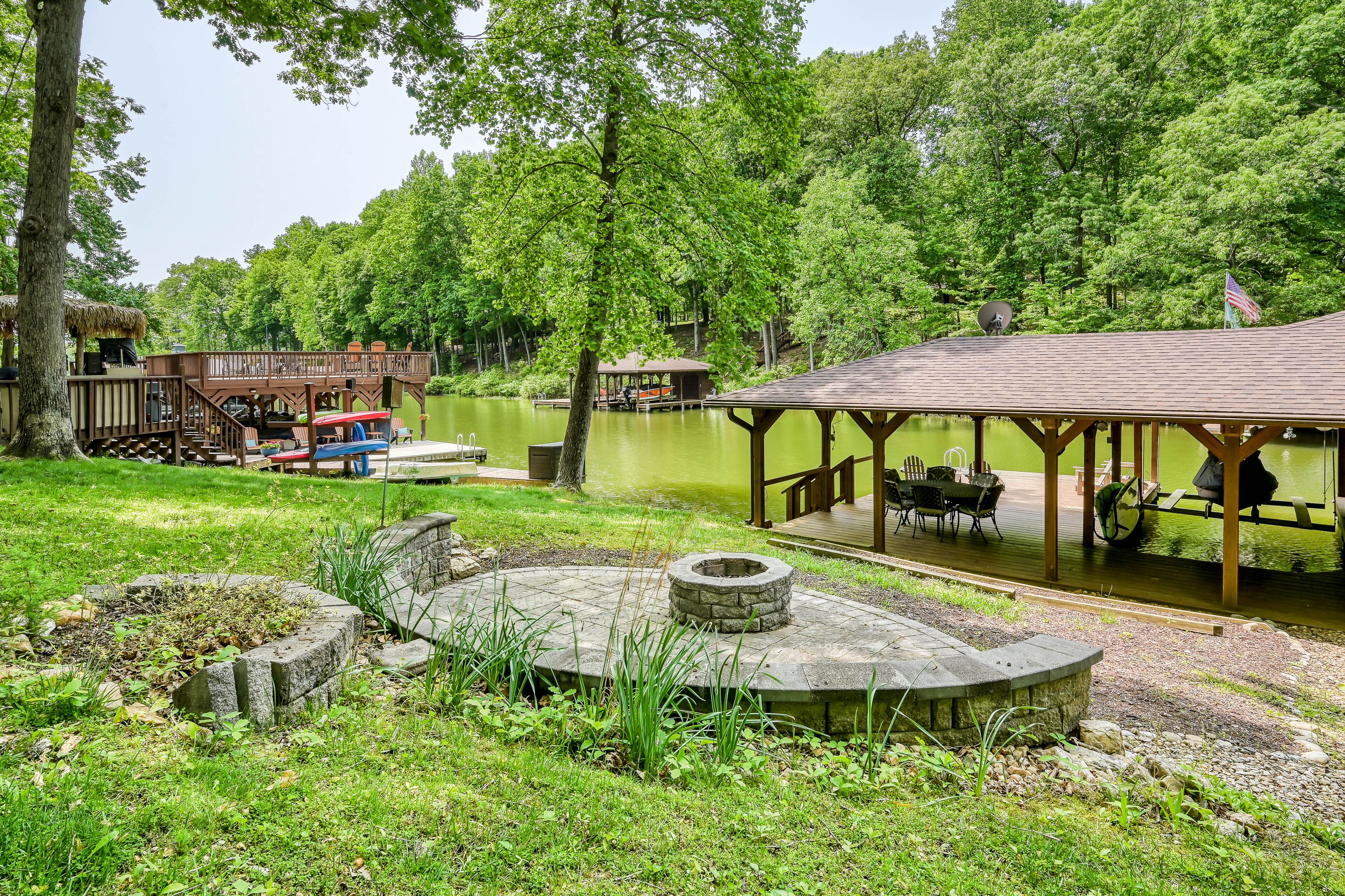 Un parque junto al lago con una terraza de madera, mesas de picnic y un hogar a leña de piedra.