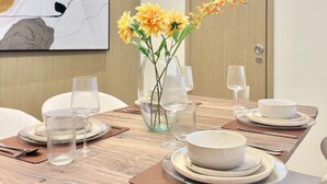 Une salle à manger moderne avec une longue table, de la verrerie transparente et un vase rempli de fleurs jaunes.