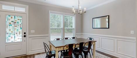A dining room with a wooden table and chairs, a chandelier, a patterned rug, and a window with blinds.
