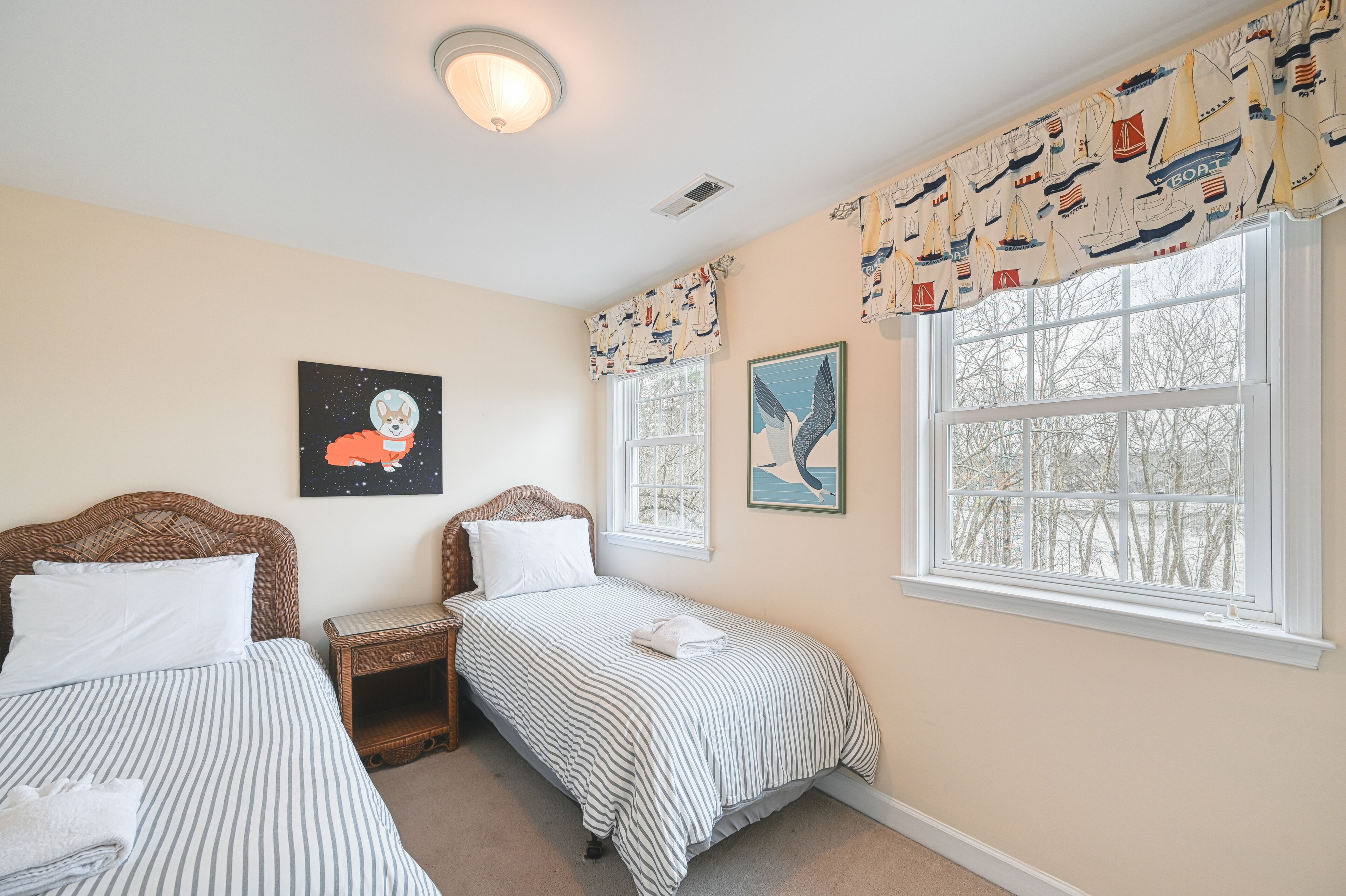 Un dormitorio con dos camas individuales, una mesita de noche, una ventana con vistas a árboles y cortinas con motivos náuticos.