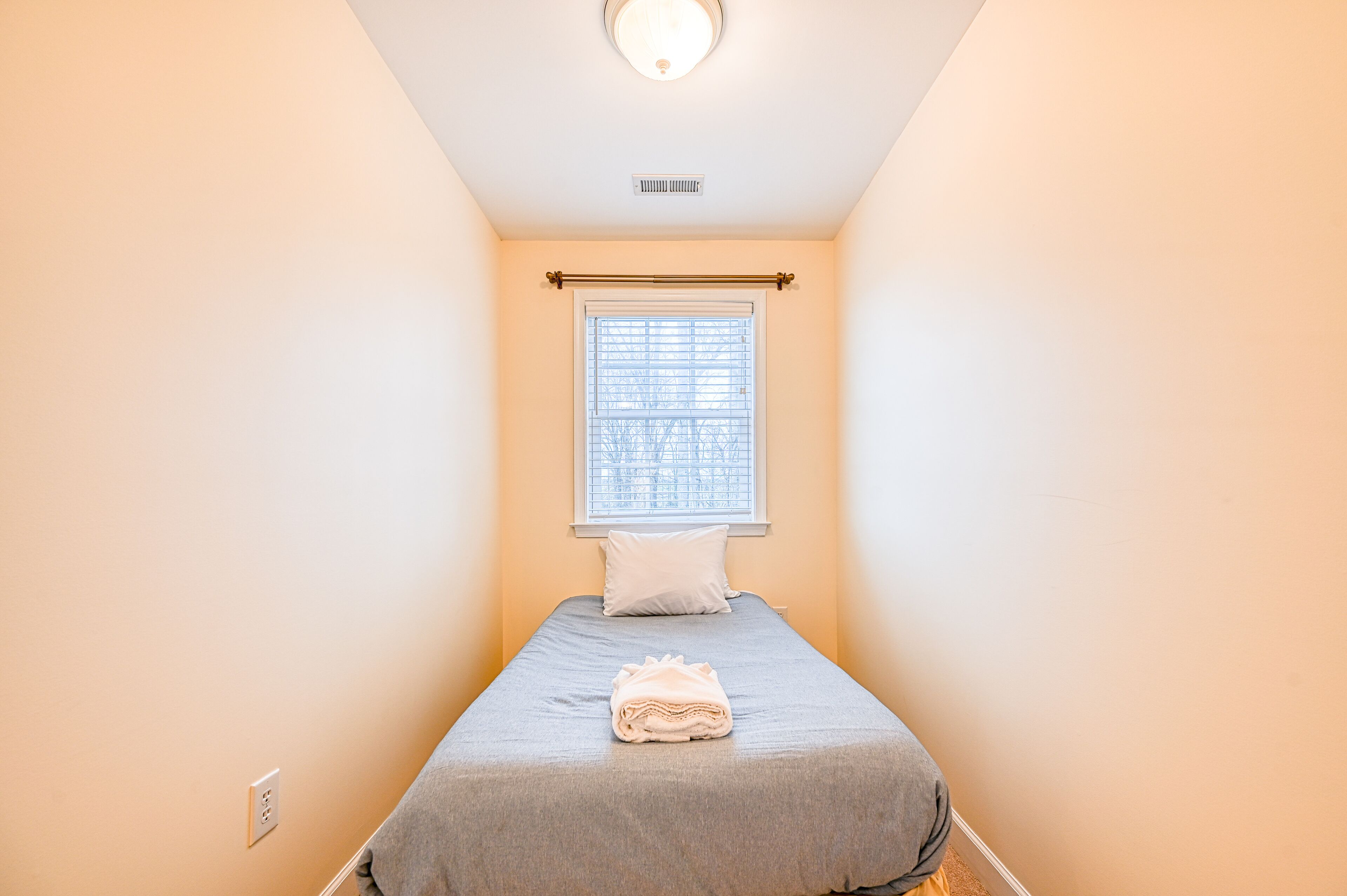 A small, neatly made bed with a grey bedspread and a folded towel, a window with blinds, and a light fixture on the ceiling.