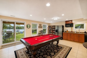 A pool table in a room with a view of a lake, a kitchen area, and a dining area with outdoor seating.