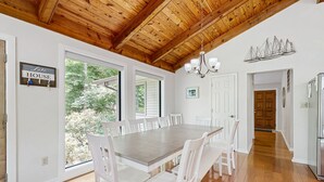 Un'ampia sala da pranzo con un grande tavolo in legno e sedie bianche, un lampadario e vista su alberi attraverso ampie finestre.