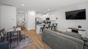 Eine moderne Küche mit weißen Schränken, eine Essecke mit Tisch und Stühlen, ein graues Sofa und ein flachbildfernseher an der Wand.