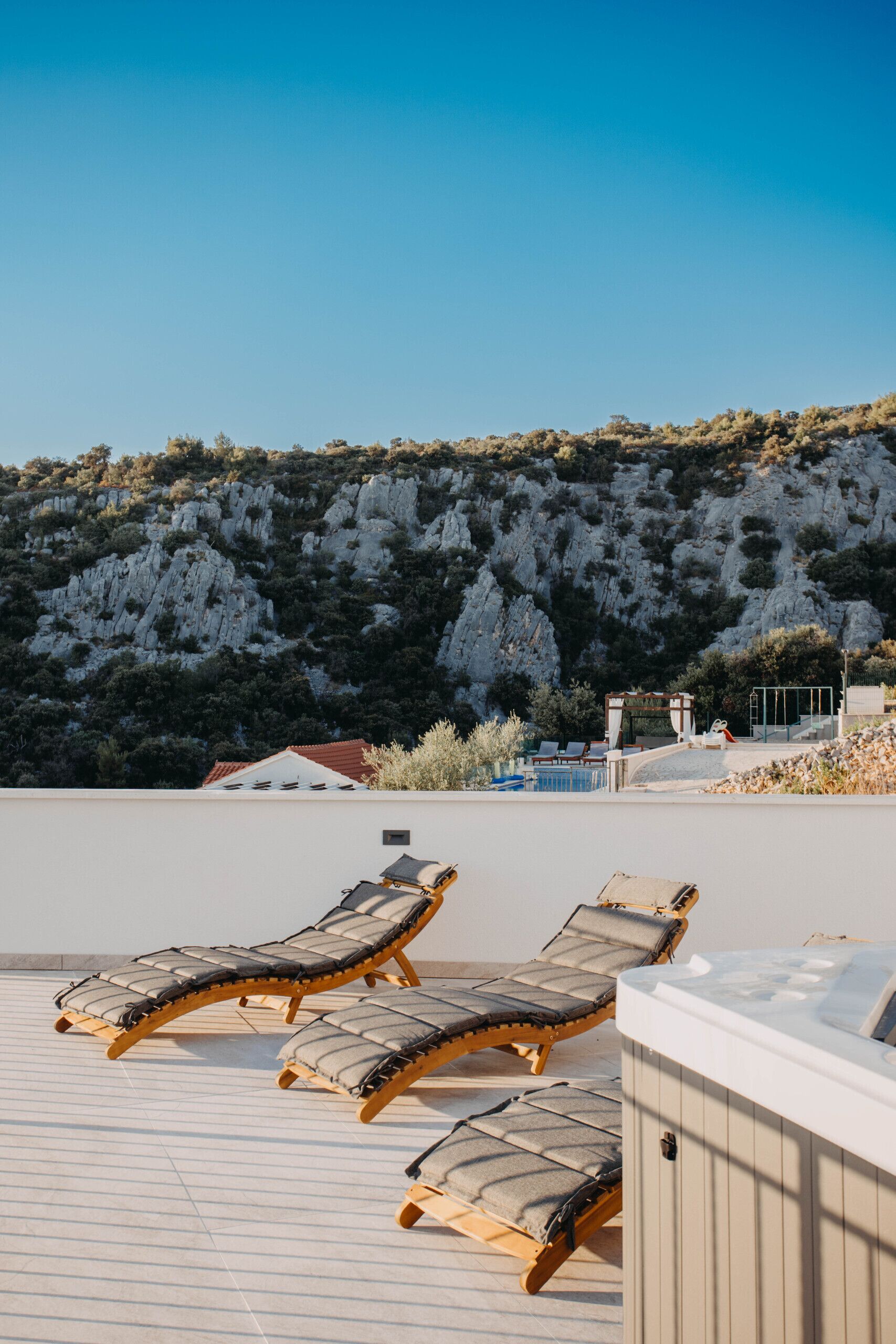A rooftop terrace with lounge chairs, a hot tub, and a view of a mountainous landscape.