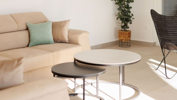 A modern living room with a beige sofa, a round coffee table, and a dark chair. There is a potted plant and a wall-mounted light fixture. - Villa La Fortezza With sea View (Marina)