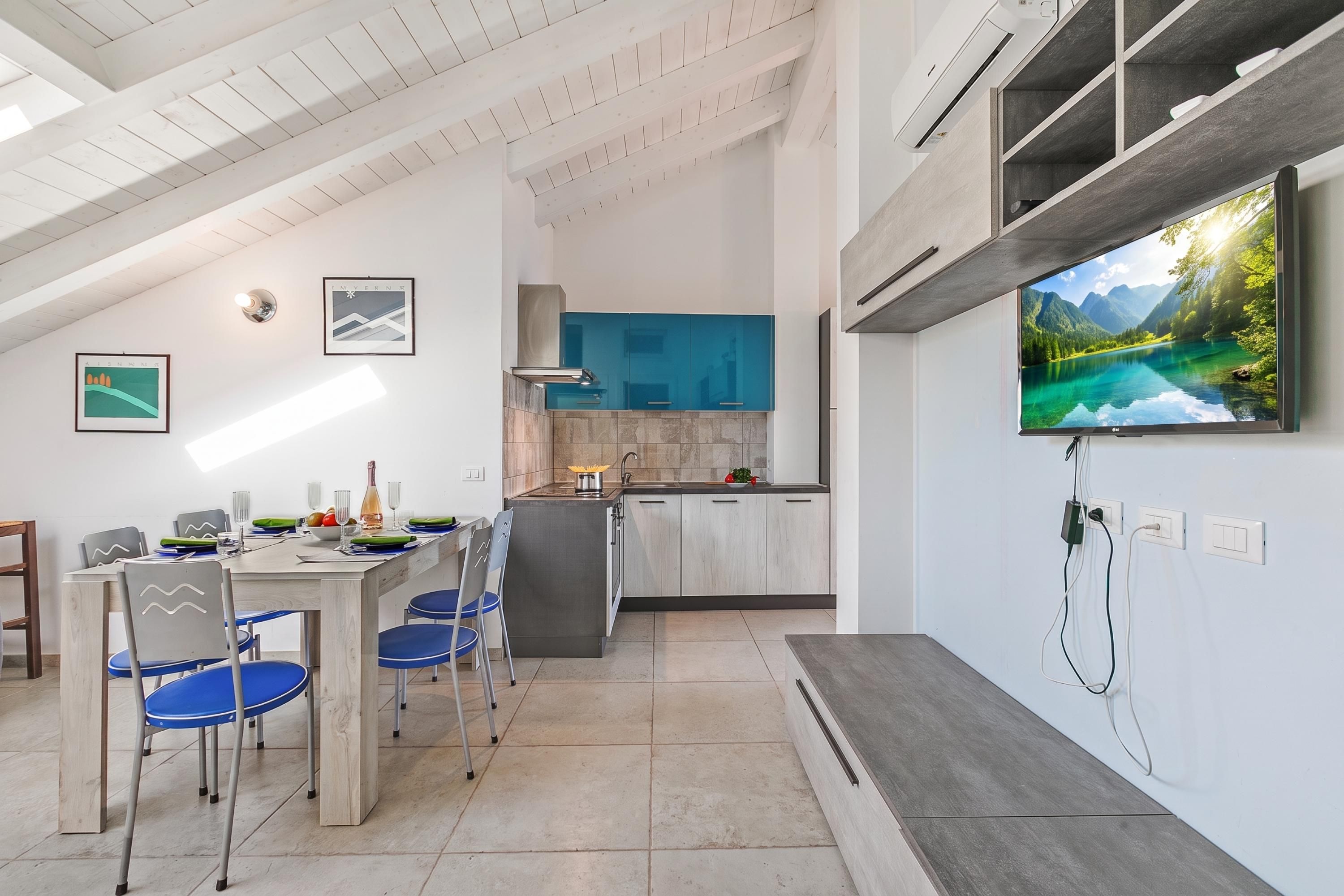 Una cucina moderna con piano cottura integrato, un'area pranzo con sedie blu e un grande televisore a schermo piatto montato a parete.