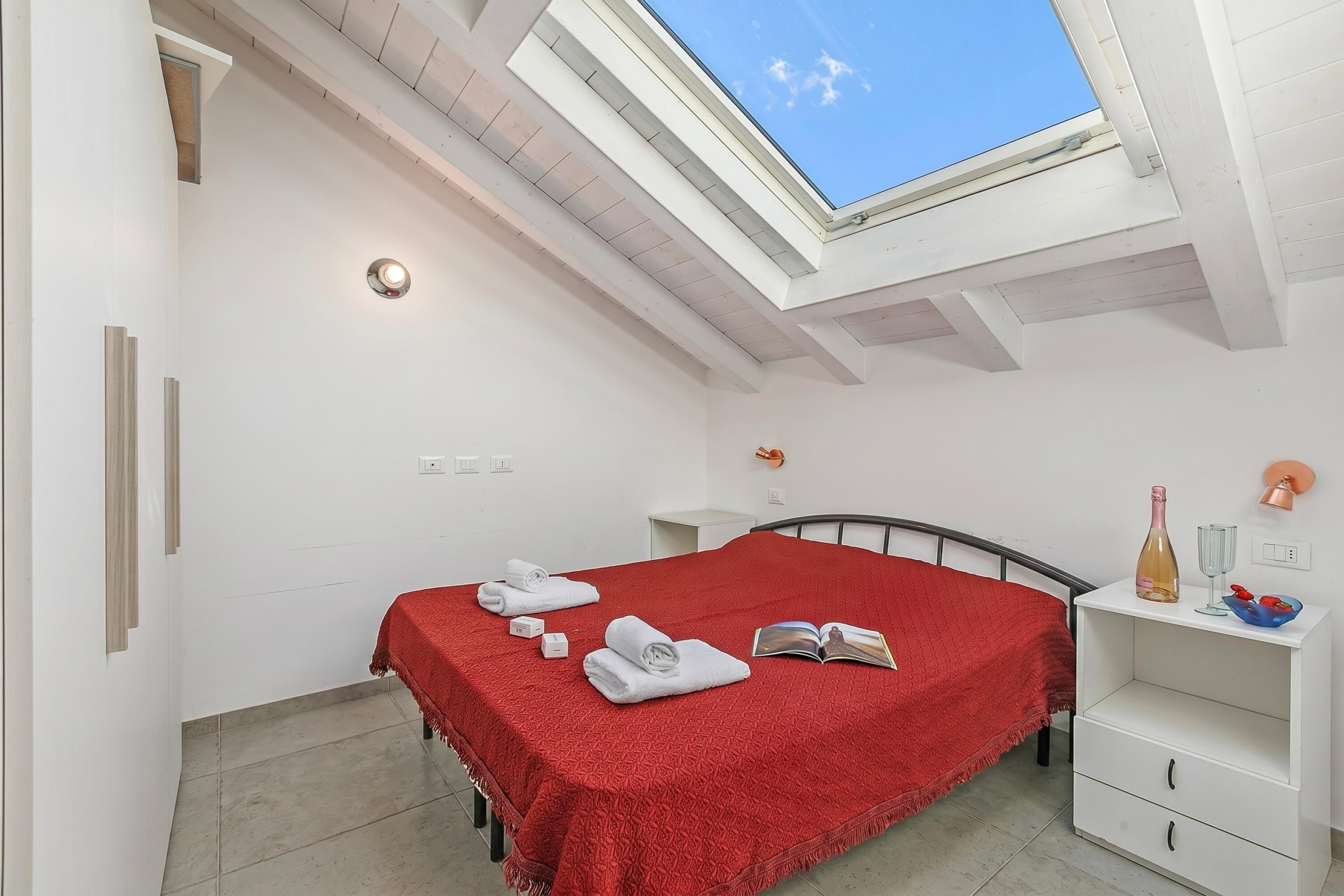 Ein Schlafzimmer mit Dachfenster, einem Bett mit roter Decke, einem Nachttisch mit Flasche und Gläsern sowie einem weißen Nachtkästchen.
