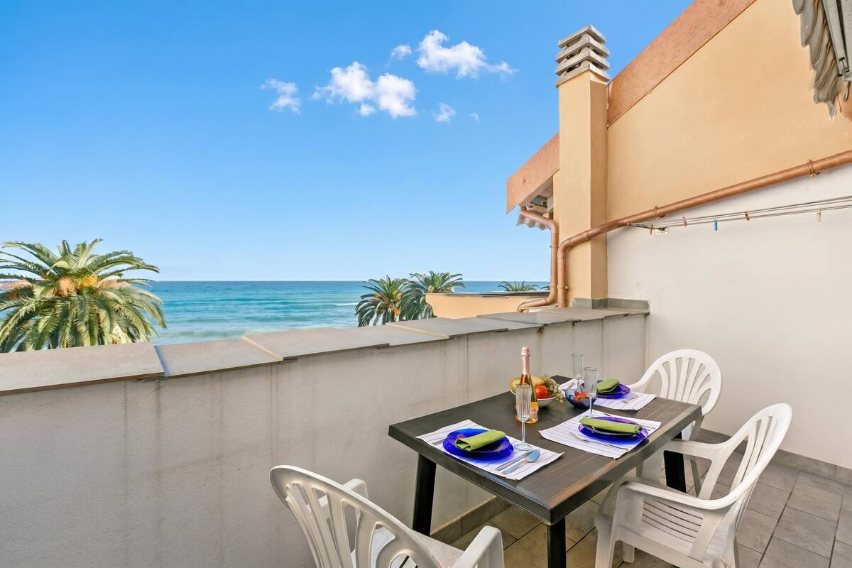 A balcony with a table set for two, overlooking a beach with palm trees and the sea.