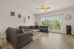 A living room with a ceiling fan, large windows, and a view of greenery outside.