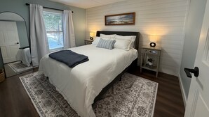 Un dormitorio con cama, lámparas de noche, ventana con cortinas, puerta y un cuadro colgado en la pared.