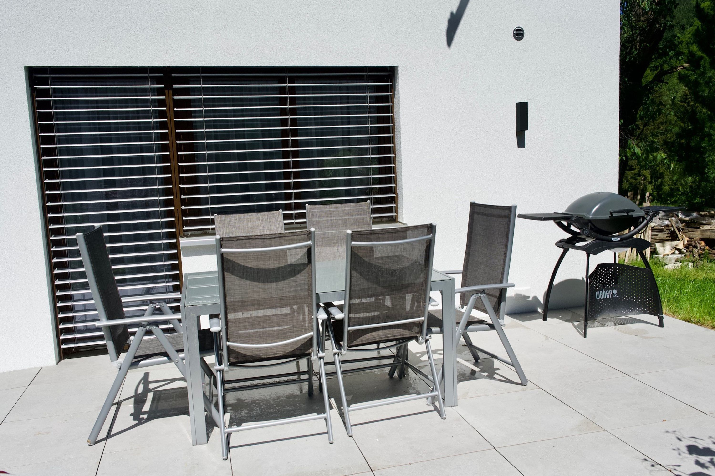 Un espacio exterior para comer con una mesa, sillas, una parrilla y una ventana con persianas.