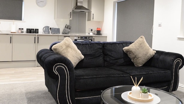Een moderne woonkamer met een zwarte bank, twee beige kussens, een glazen salontafel met taart en kaarsen, en een keuken op de achtergrond.