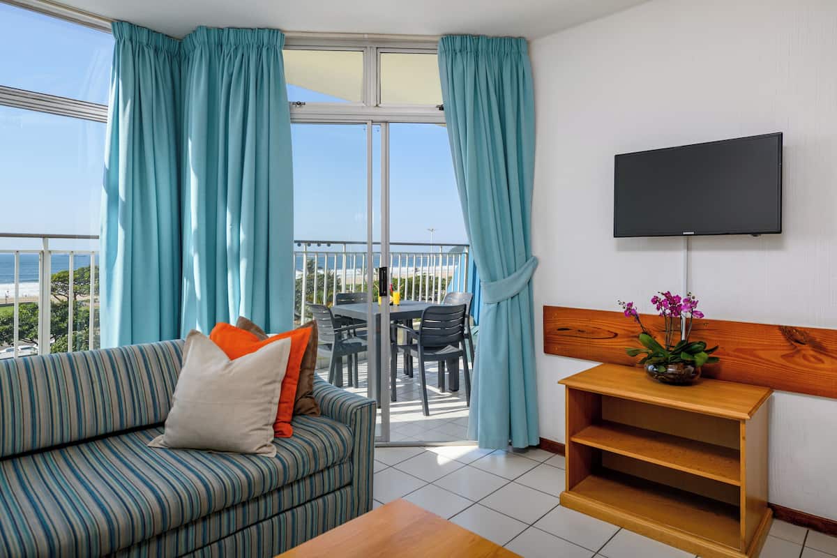 A balcony with a table and chairs, a sofa with cushions, a TV mounted on the wall, and a wooden side table with a plant.