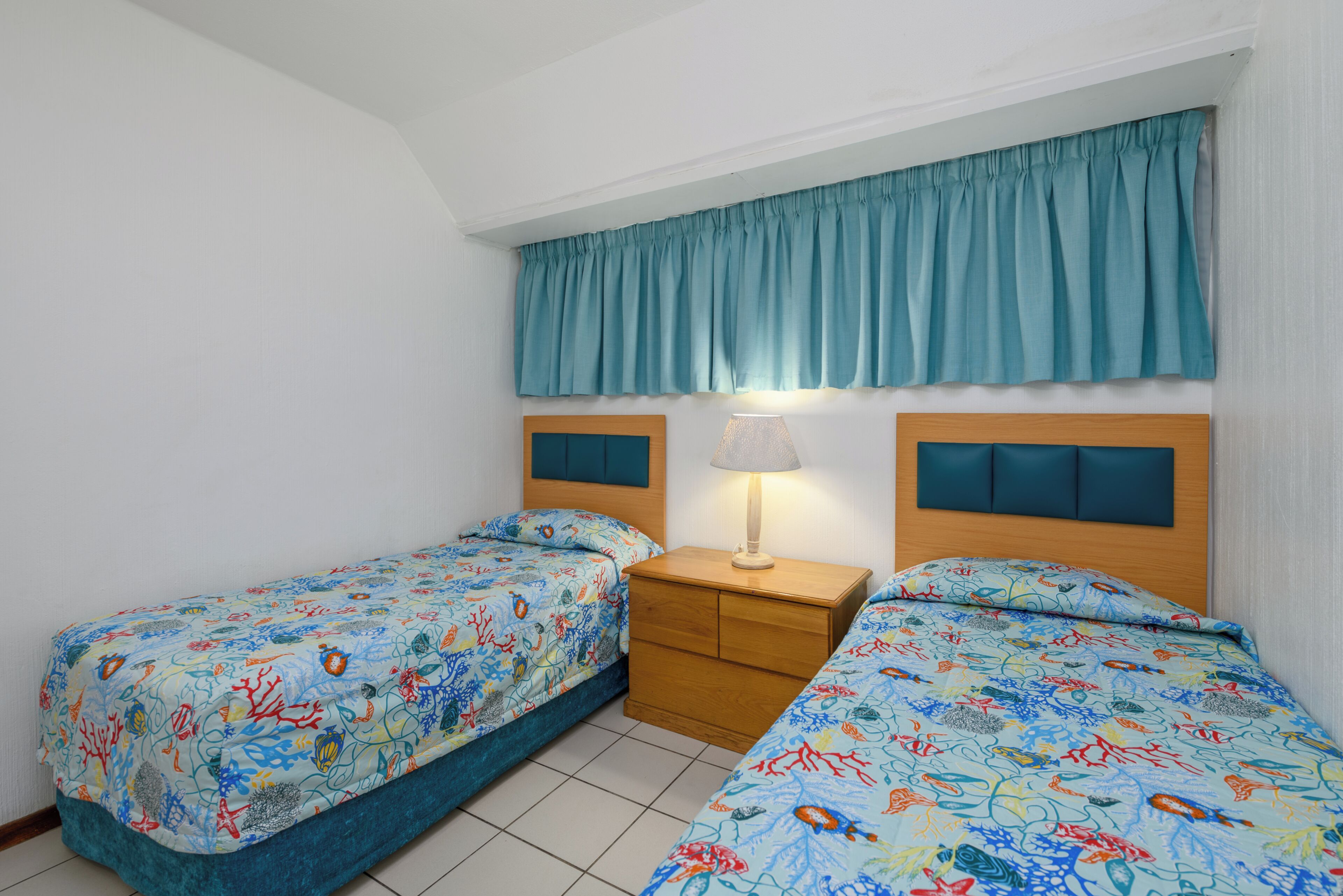 Un dormitorio con dos camas individuales, una mesita de noche con lámpara y una ventana con cortina verde azulado.