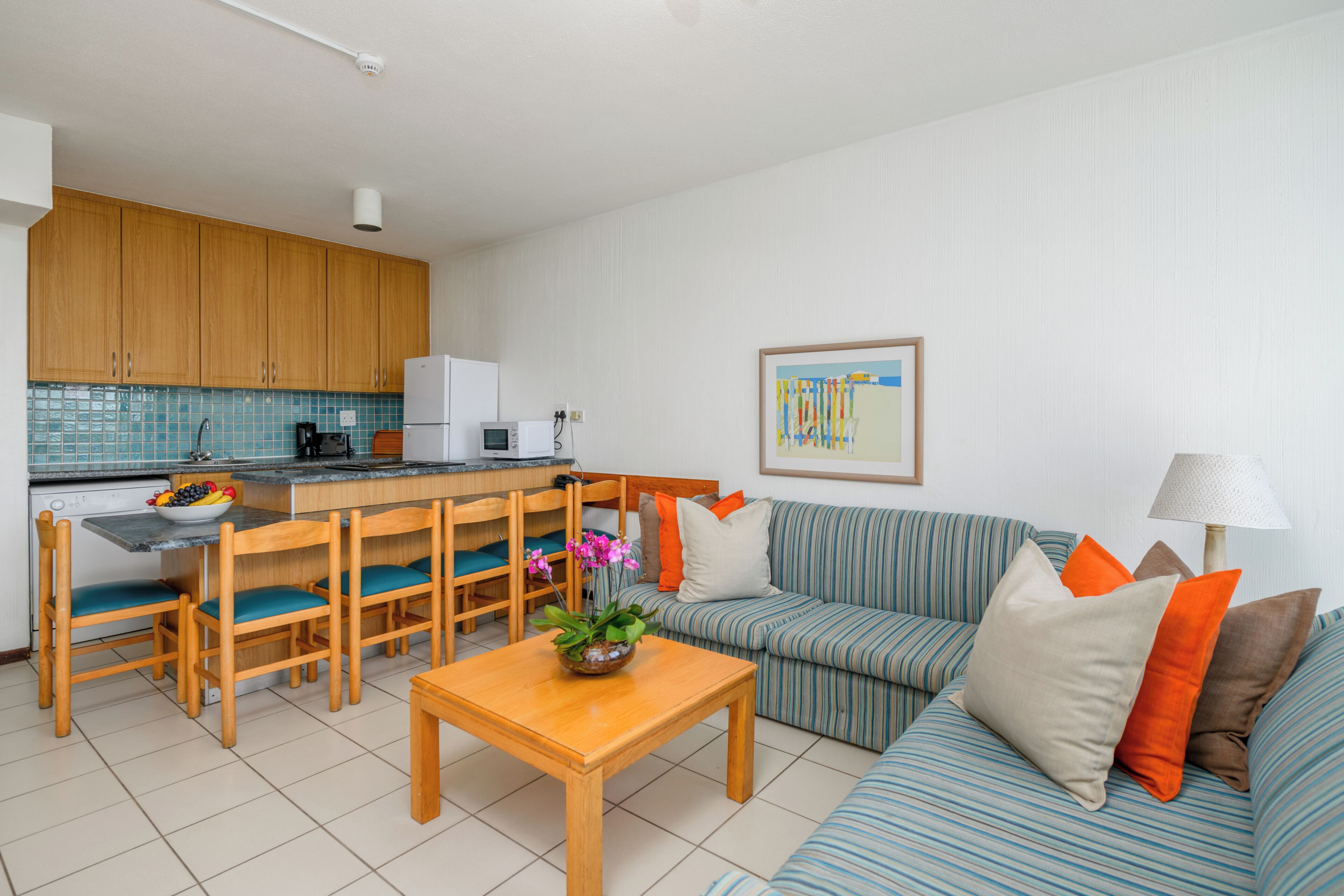 Una cocina moderna con armarios de madera, una zona de comedor con sillas y una mesa, un sofá a rayas con cojines, y un cuadro enmarcado en la pared.
