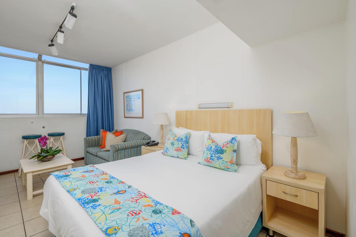 A hotel room with a bed, a nightstand, a lamp, a chair, a table with a plant, and a window with curtains.