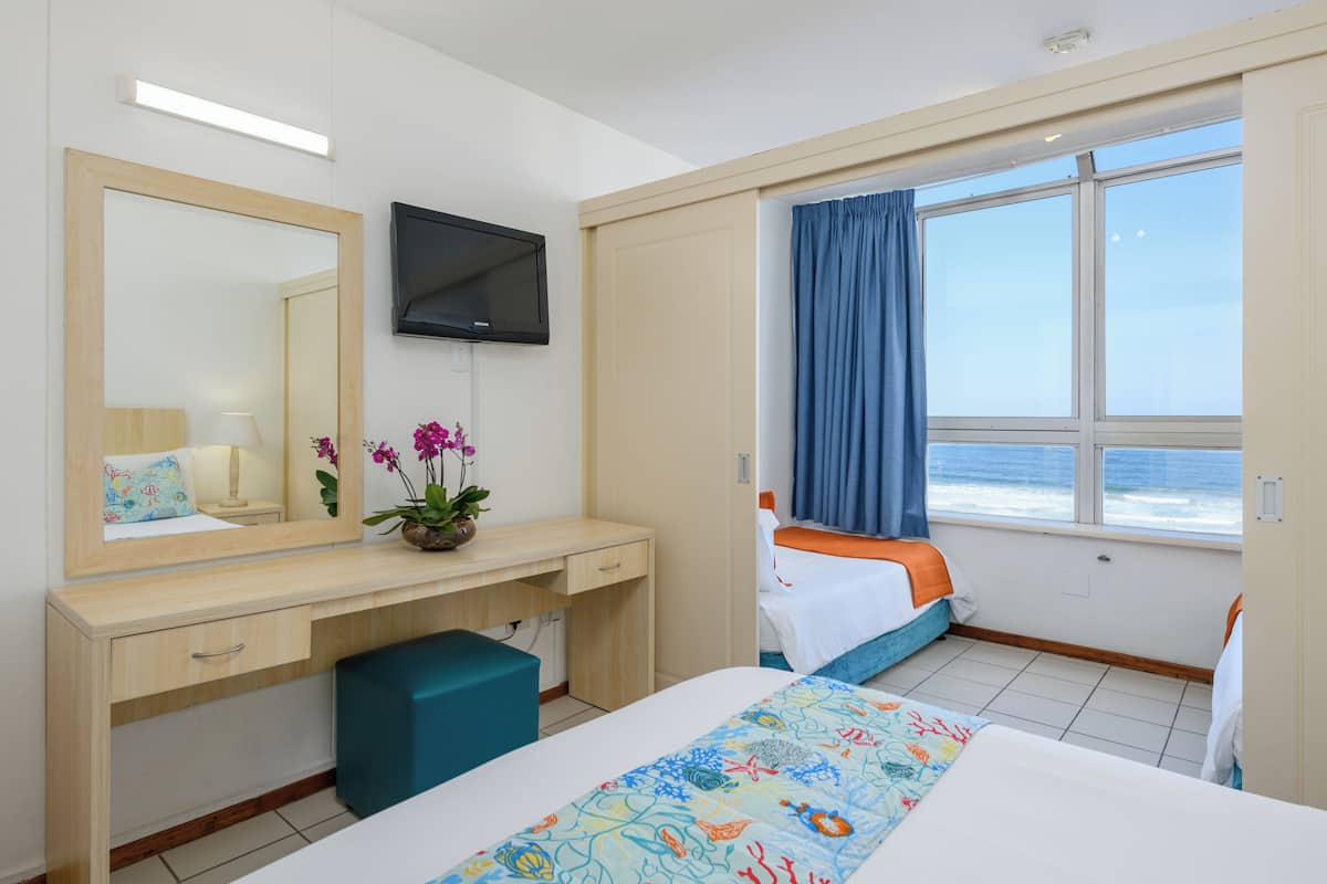 A hotel room with a bed, a desk with a TV, a window with a view of the sea, and a bathroom.
