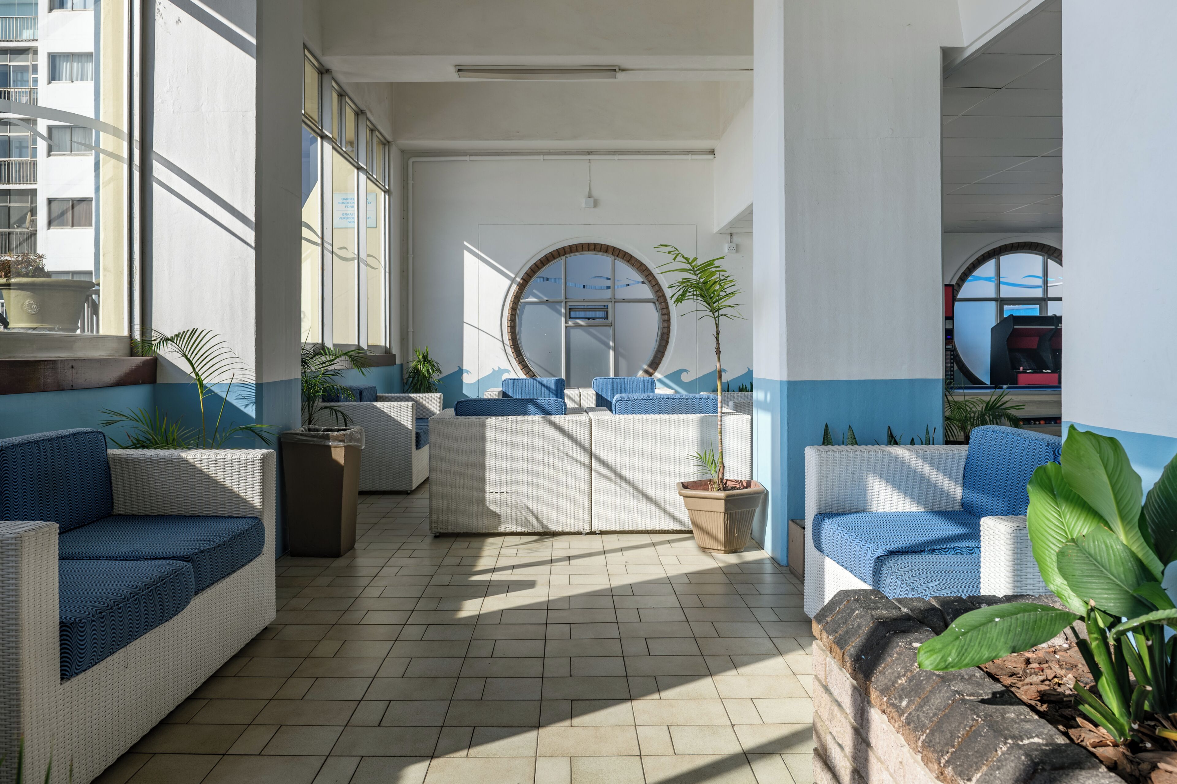 Espaço interior iluminado pelo sol, com móveis de vime, plantas em vasos e uma grande janela.