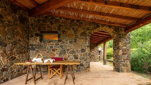 A stone-walled outdoor area with a wooden table and chairs, a stone archway, and a view of greenery.