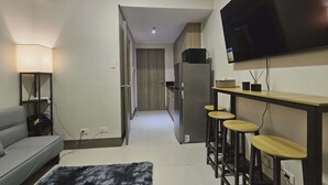 A modern living room with a sofa, a TV mounted on the wall, a small kitchen area, and a hallway leading to another room. - Casa Rogelio at Fame Residences Tower 4 (Mandaluyong)