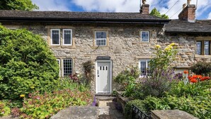 Une petite maison en pierre avec une porte blanche, de nombreuses fenêtres et un jardin où poussent différentes sortes de fleurs.