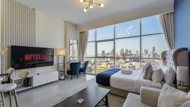 Ein modernes Hotelzimmer mit einem großen Bildschirm, auf dem 'NETFLIX' angezeigt wird, Blick auf die Stadt durch große Fenster, einem bequemen Bett und einer Sitzecke mit Couchtisch.
