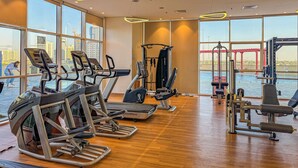 Ein Fitnessstudio mit Laufbändern, Crosstrainern und Gewichtsmaschinen. Große Fenster bieten Blick auf eine Brücke und Gebäude.
