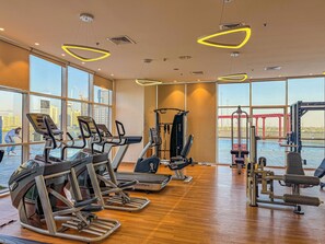 Un gimnasio con cintas de correr, elípticas y equipo de pesas. Amplias ventanas ofrecen vistas de un puente y edificios.