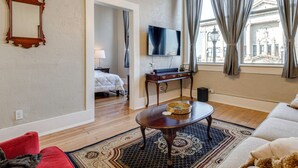 A hotel room with a wooden floor, a large mirror, a flat-screen TV, a dining table with a basket, a sofa, and a view of a building through the window. - Historic Lebanon Gem: Central Walkable Location! (Lebanon)
