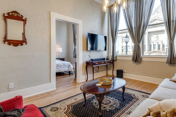 Habitación de hotel con piso de madera, un espejo grande, televisor de pantalla plana, una mesa de comedor con un cesto, un sofá y vista a un edificio a través de la ventana.