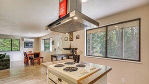 A modern kitchen with a central island, a dining area with a table and chairs, and large windows with a forest view. - Creative Escape! Eagle's Nest Hideaway (Cottage Grove)