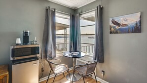 A compact room with a kitchenette, a small round table with two chairs, a painting of a bird, and a view of a body of water through large windows. - Main-channel Views for Lake Ozark Shootout Weekend (Lake Ozark)
