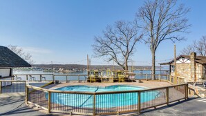 A swimming pool with a wooden deck and metal railing, surrounded by a fence, with a view of a lake and buildings in the background. - Main-channel Views for Lake Ozark Shootout Weekend (Lake Ozark)