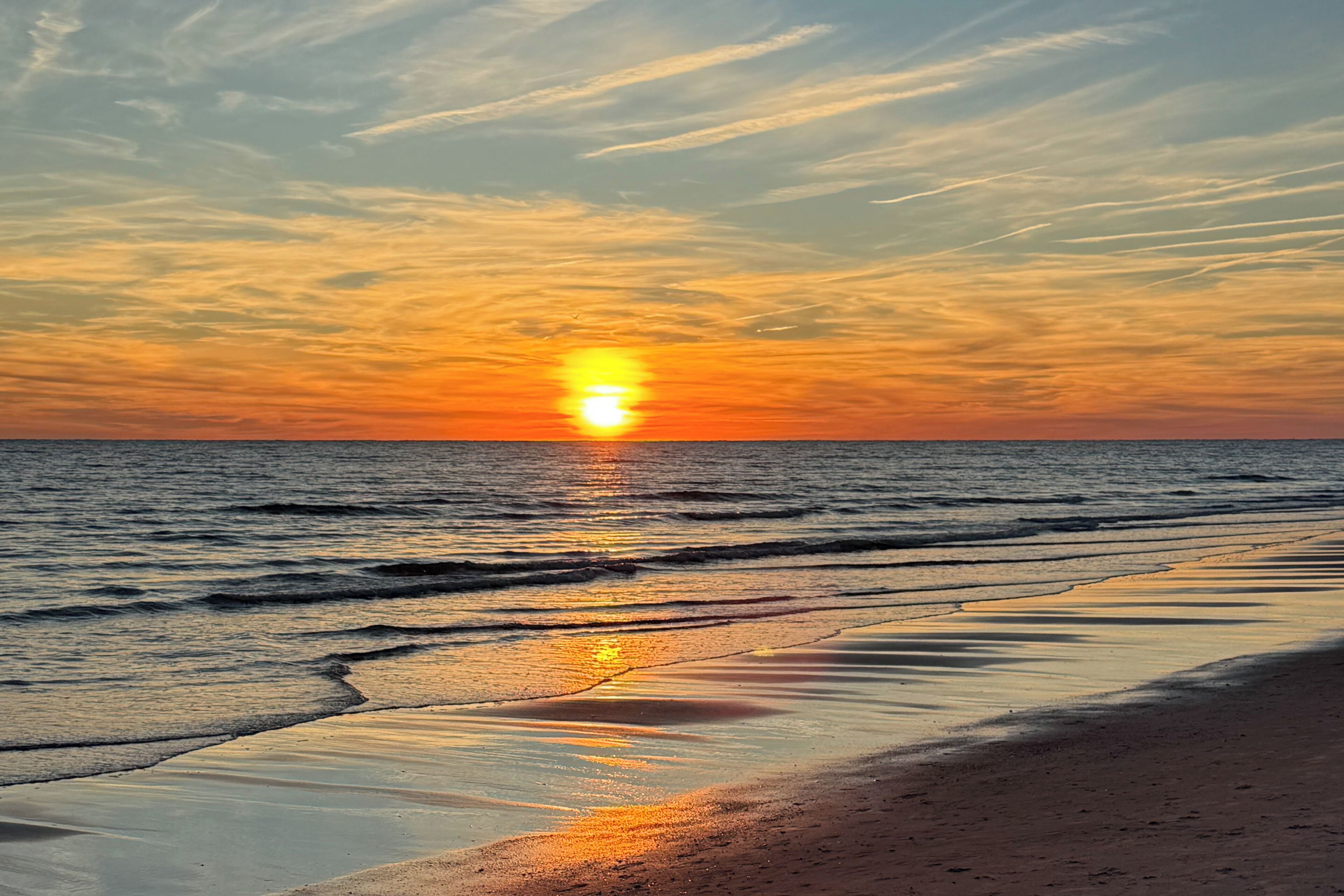A beach with the sun setting over the horizon, calm water, and a clear sky.