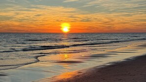 A beach with the sun setting over the horizon, calm water, and a clear sky. - Spacious Coastal Gem on Oak Island 100 Yds to Sand (Oak Island)