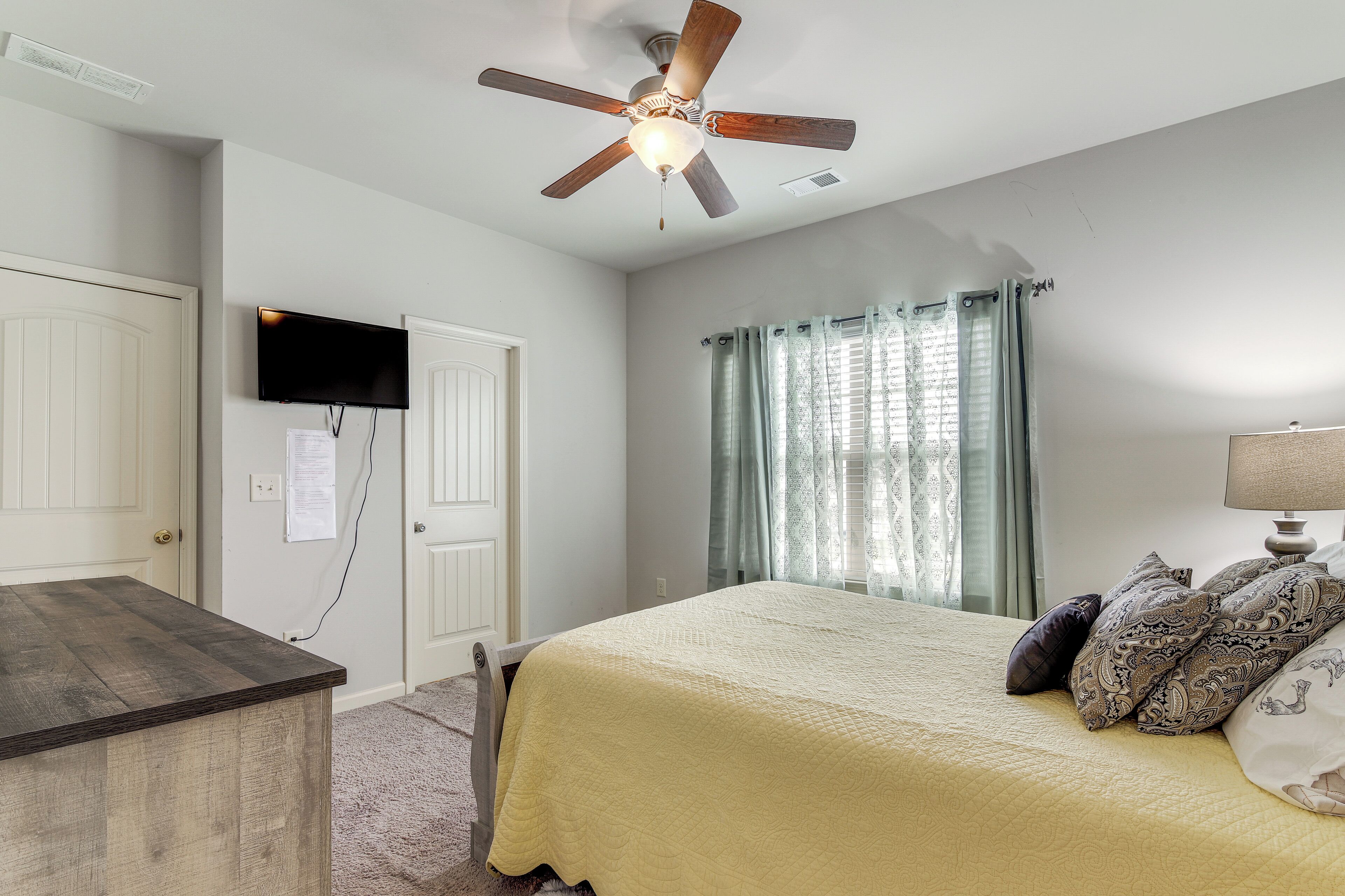 A bedroom with a bed, a ceiling fan, a TV mounted on the wall, and a window with curtains.