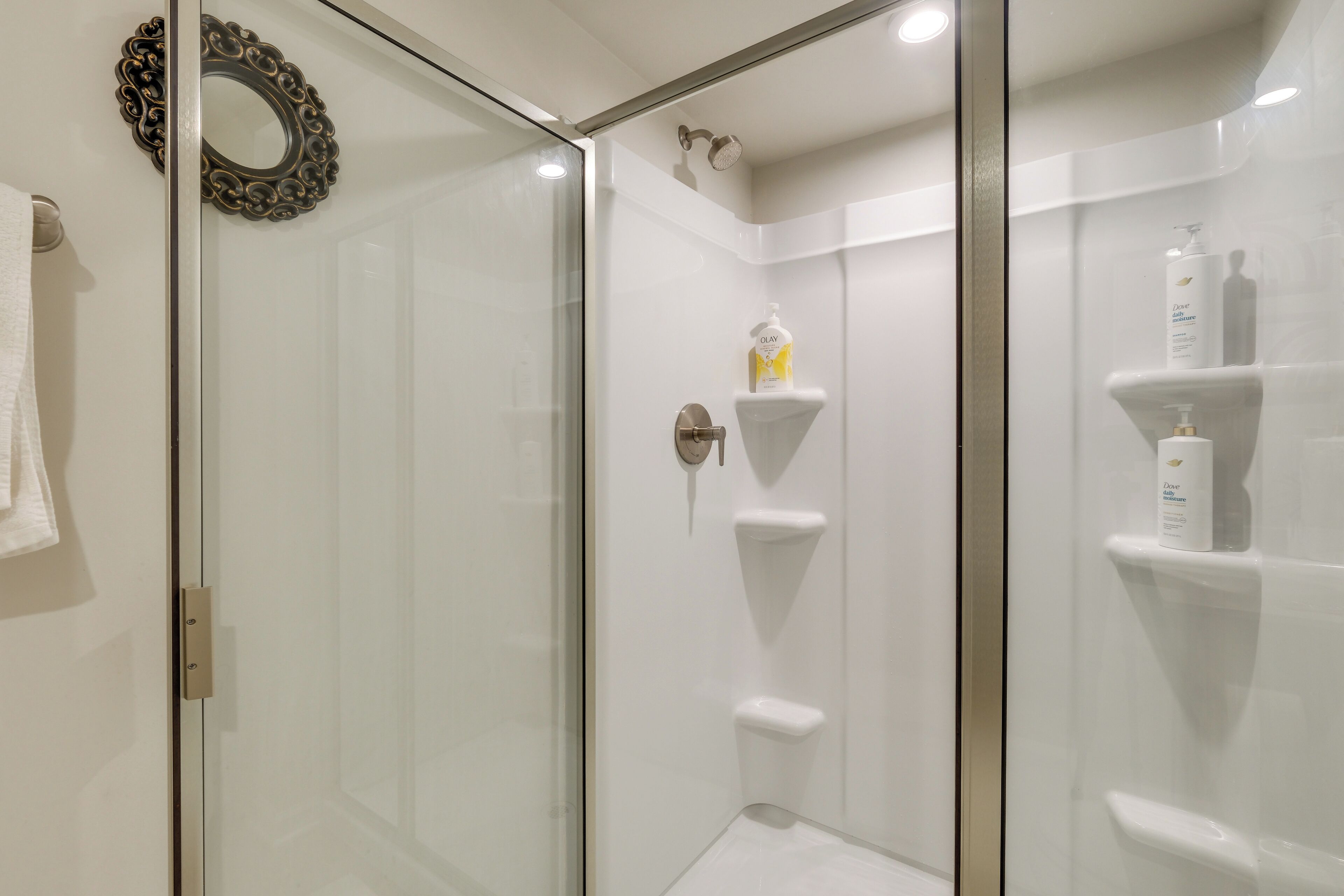 A glass-enclosed shower with a white tiled wall, a decorative mirror, and toiletries on a shelf.