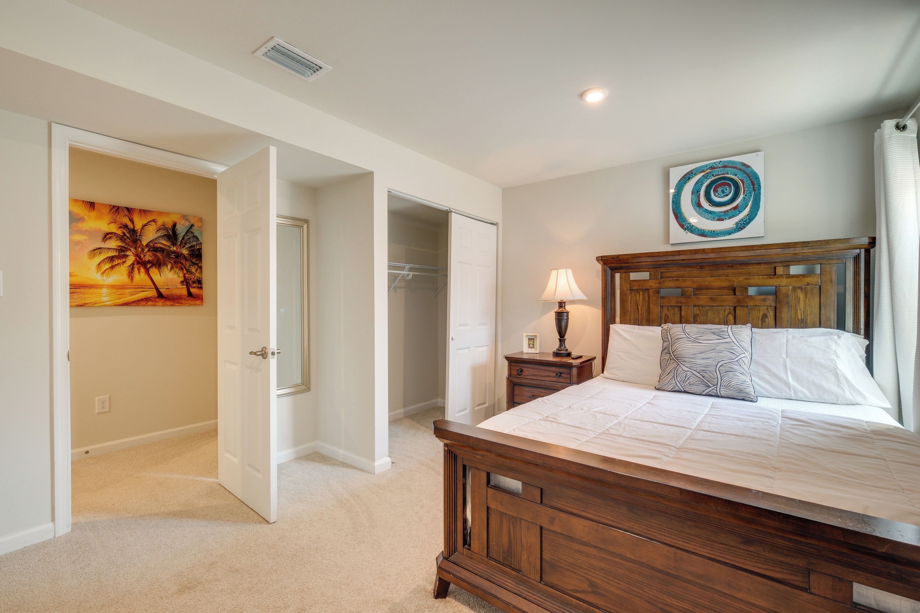 A bedroom with a wooden bed, a nightstand, a lamp, a painting of a sunset, and a closet.