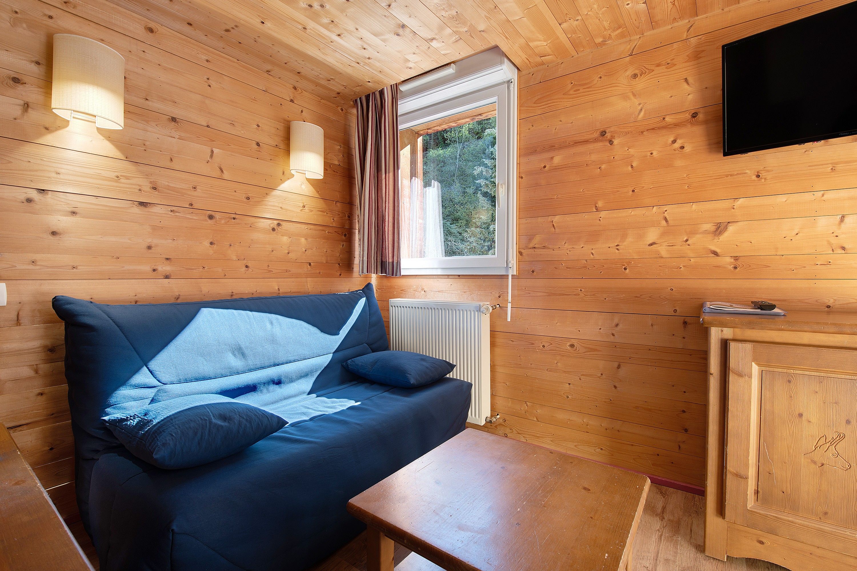 A wooden cabin interior with a sofa, a table, a radiator, and a television mounted on the wall.