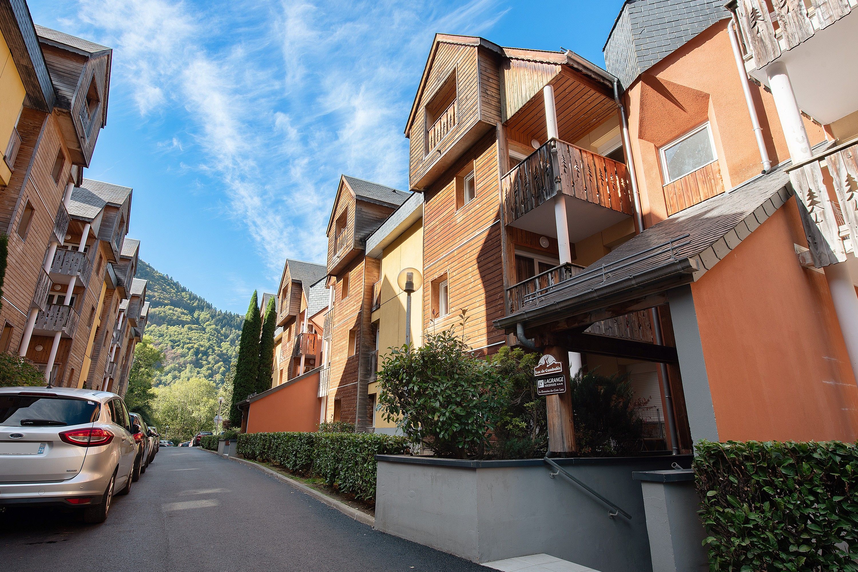 A row of multi-story wooden buildings with balconies, a clear sky, and a car parked on the street.