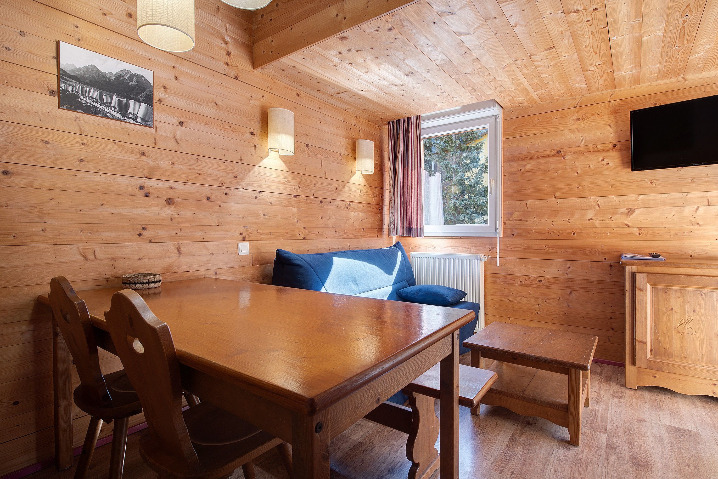 A wooden cabin interior with a dining table, chairs, a sofa, a television, and a window with curtains.