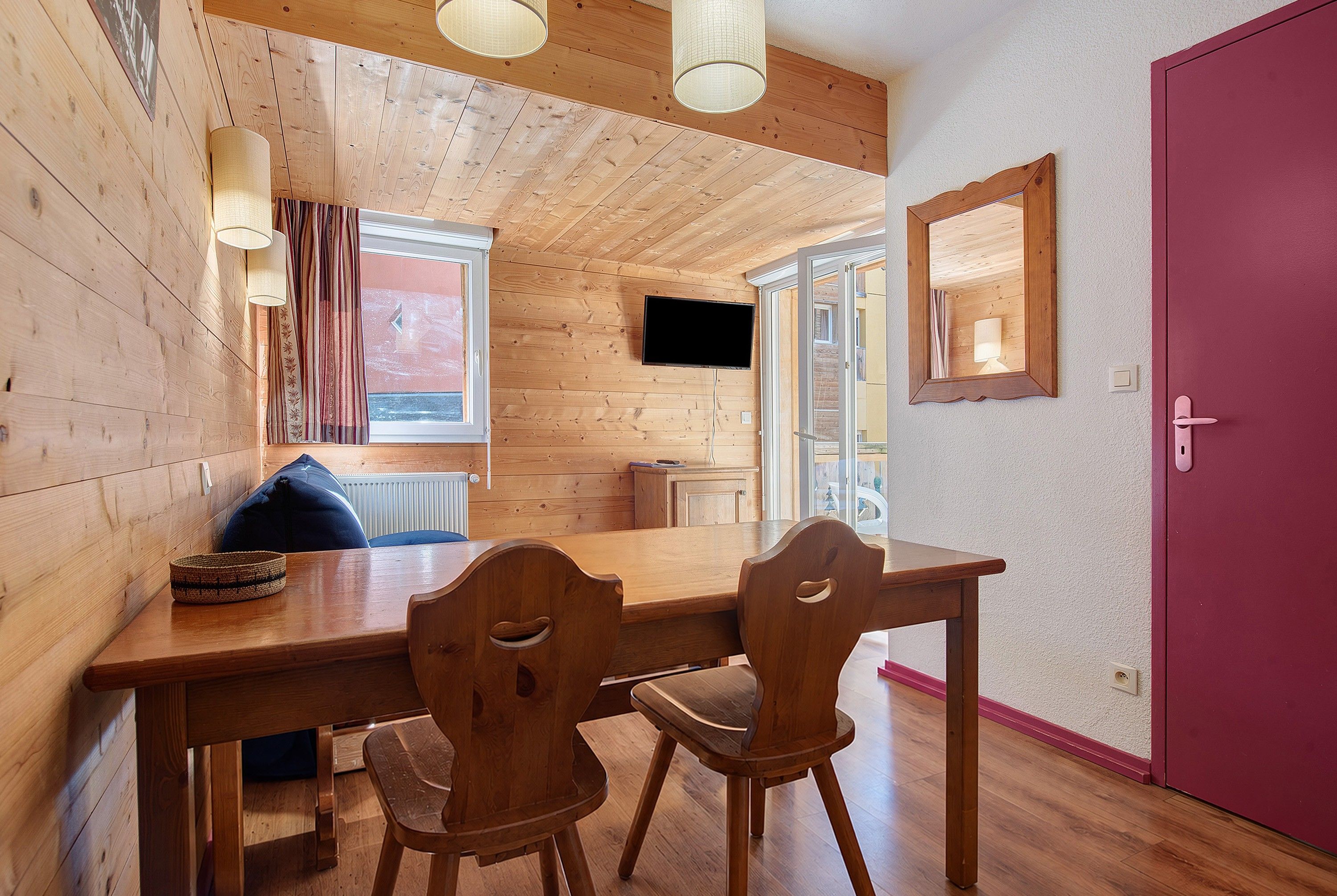 A wooden interior with a dining table, chairs, a television mounted on the wall, and a door.