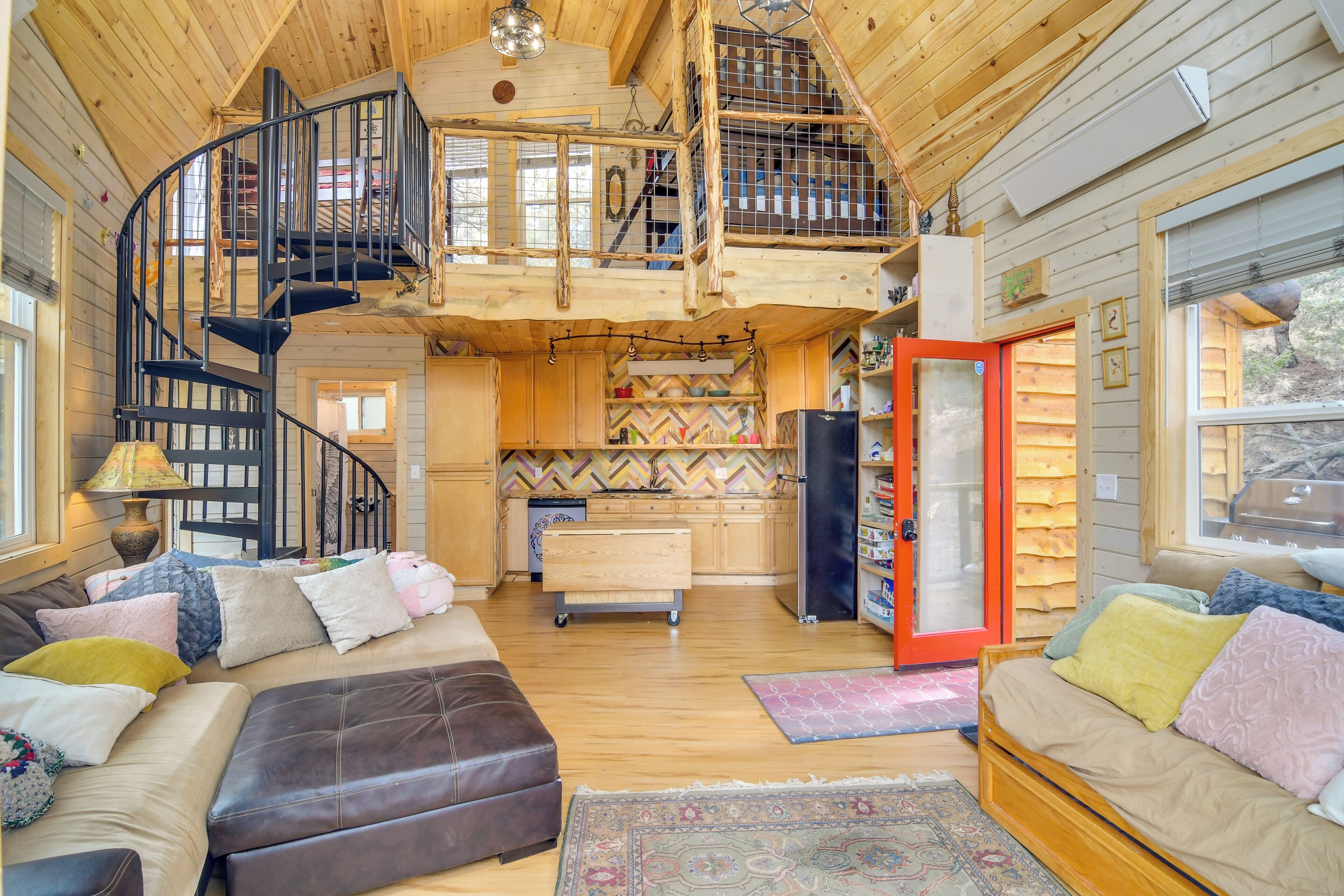 A cabin interior with a loft bedroom, a kitchen, and a living area with a sofa and ottoman.