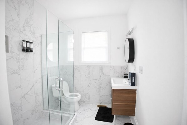 A modern bathroom with a glass shower enclosure, a white toilet, a wooden vanity, and a marble backsplash.
