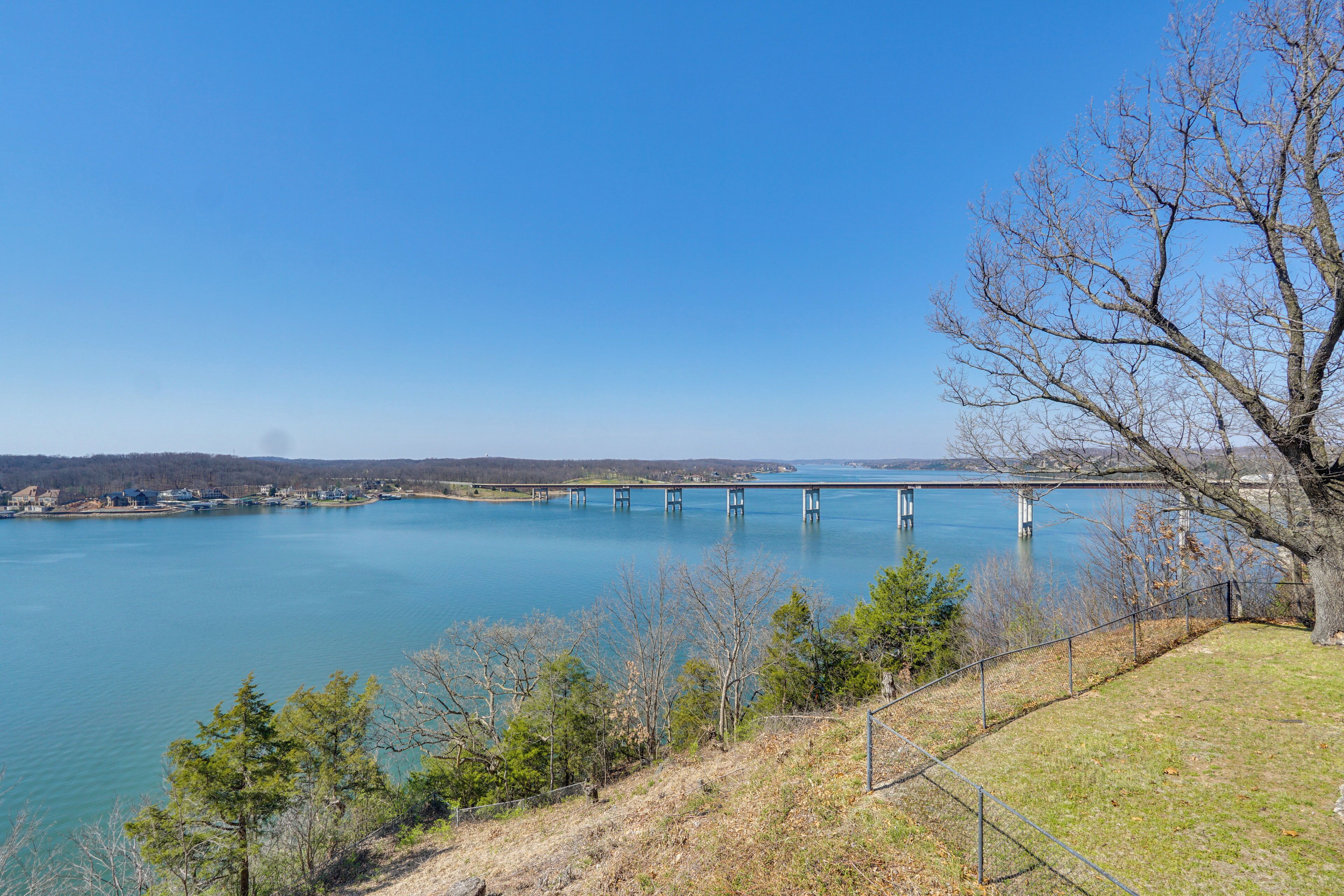 A bridge over a calm lake with a clear sky, surrounded by trees and a fenced area.