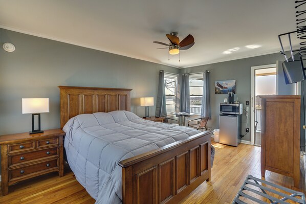 A bedroom with a wooden bed, two bedside tables with lamps, a ceiling fan, a view through a window, and a kitchenette area. - Bagnell Strip Studio for Shootout Weekend Fun (Lake Ozark)