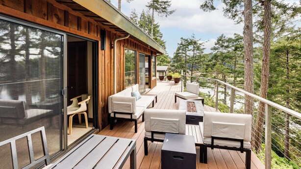 Terraza de madera con mobiliario exterior, puerta de vidrio y vista a un área boscosa.