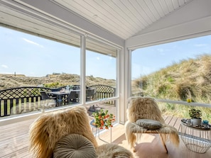 A sunlit room with a view of dunes, a wooden deck, and a dining area with outdoor furniture. - 4 Star Holiday Home in Saltum (Saltum)